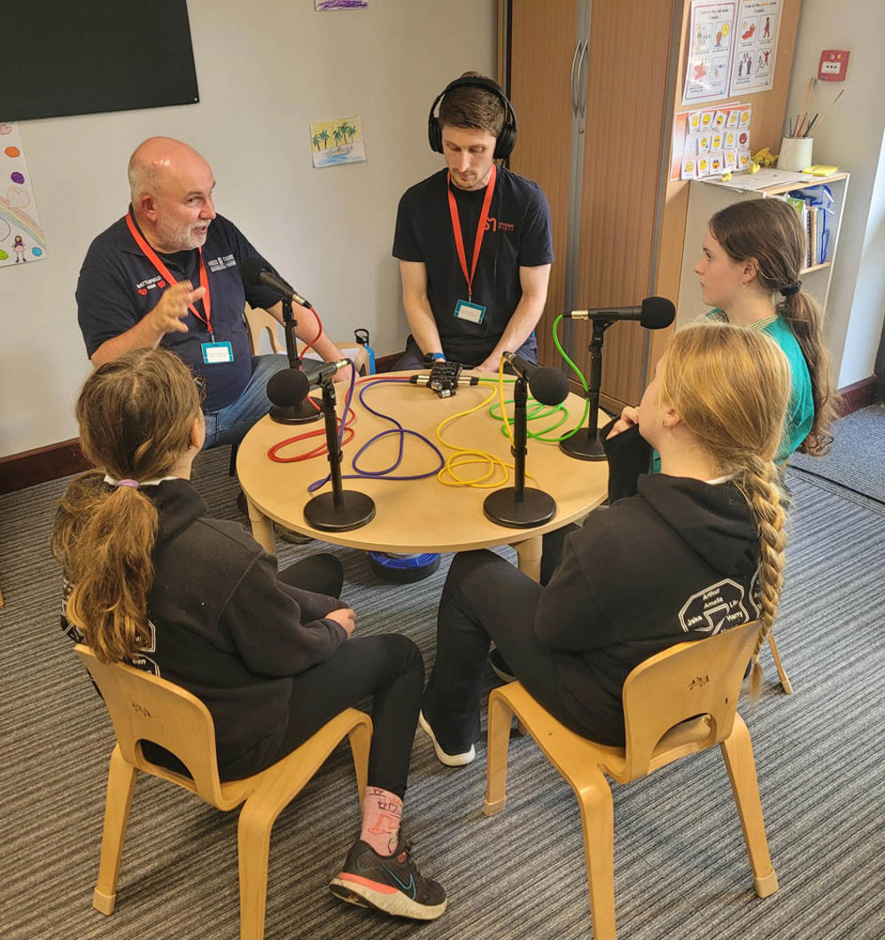 Group of schoolchildren sat around a circular wooden table interviewing members of the public. Big black microphones are placed on the table