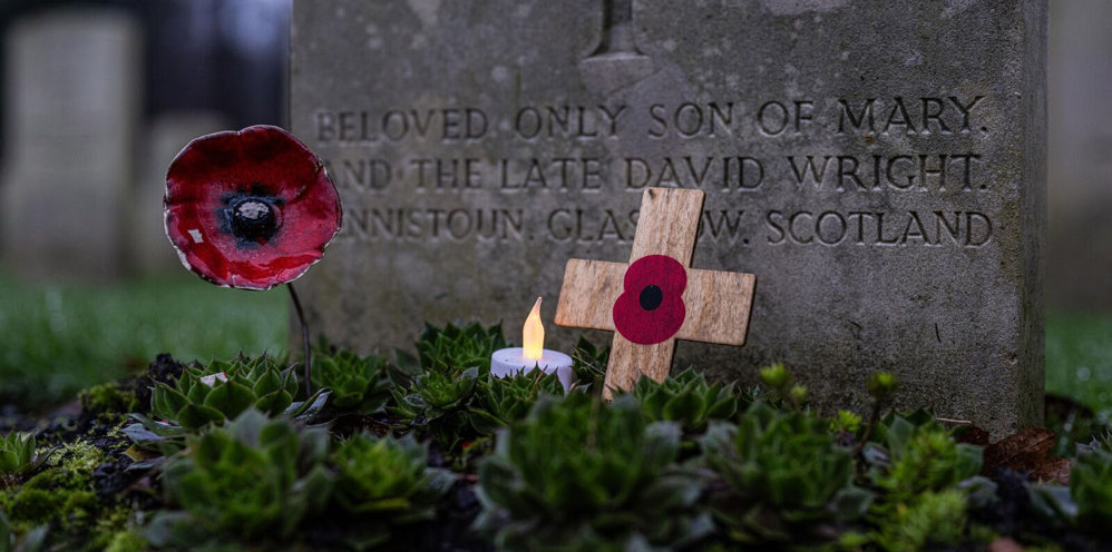 Close up of a ceramic poppy, a small wooden cross with a painted red poppy on, and a small candle at the base of a CWGC headstone.