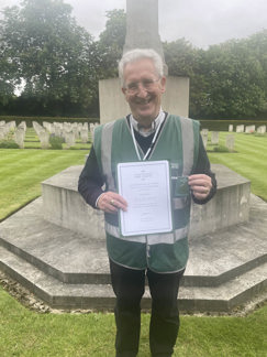 Man in hi-vis vest poses with a certificate at the base of a CWGC Cross of Sacrifice
