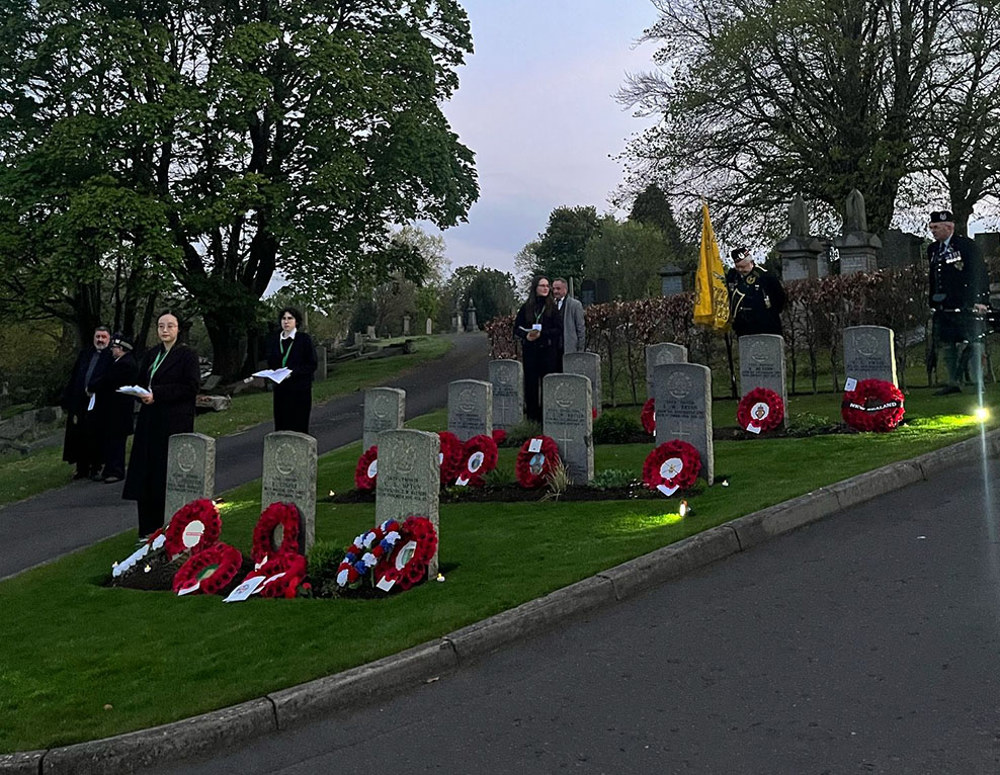 Attendees and ex-servicemen standing next to wreath-covered headstones at an ANZAC Dawn Ceremony in Glasgow Eastern Cemetery.