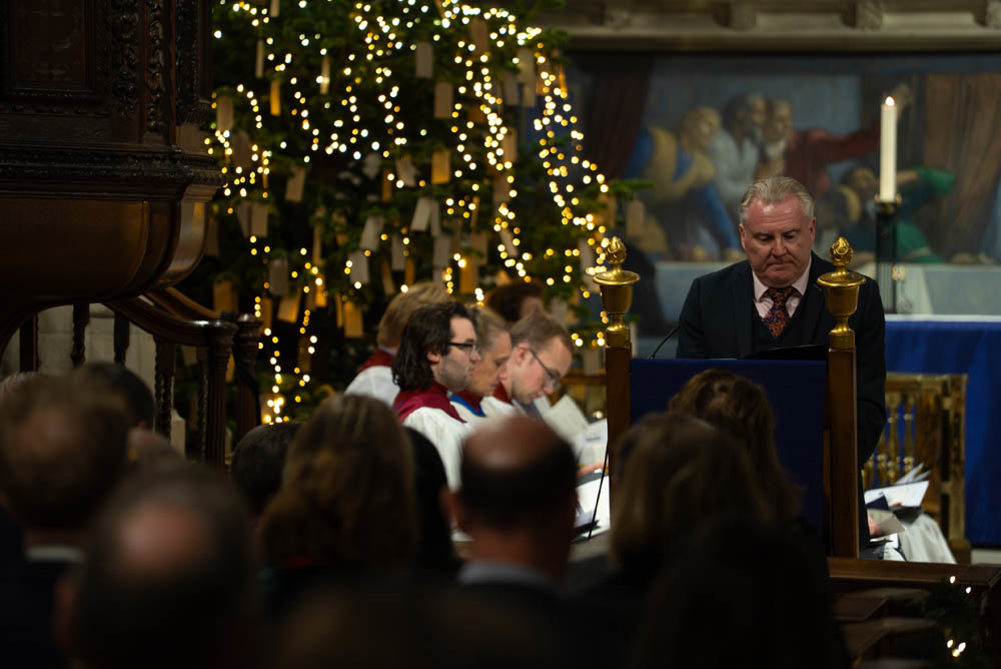 Man gives reading in a church on a lectern next to a choir and a decorated christmas tree.