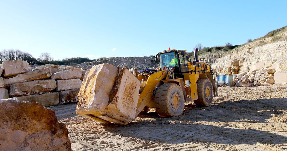A yellow digger carrying a large lump of Portland stone in its excavator.