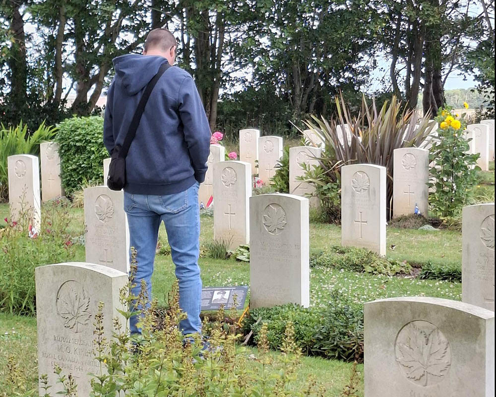 Man in a blue hoody and jeans reads a casualty story sign placed next to a war grave in a CWGC cemetery.